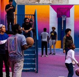 A vibrant outdoor basketball court filled with people of various ages engaged in different activities. Some are playing basketball while others are standing and chatting. The court is surrounded by brightly painted walls in shades of pink, orange, and blue. A basketball hoop is mounted on the back wall, adding to the sports theme. A few people are sitting on stairs or using phones, creating a relaxed and social atmosphere.