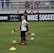 A person in sports attire stands on a soccer field, surrounded by several soccer balls and a carrier with bottles. The field is bordered by advertisements for Nike Soccer and app stores. Several people sit in bleachers in the background.