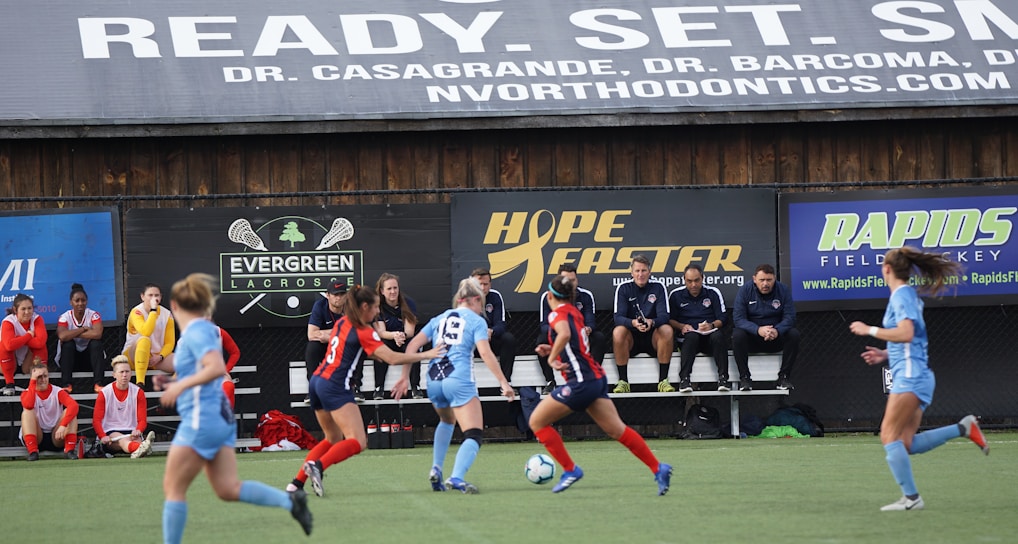 Players engage in a competitive soccer game on a field while others are seated on the bench watching attentively. The action is taking place in front of a wooden structure displaying multiple advertisements, including orthodontics, lacrosse, and field hockey.