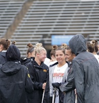 A group of people, likely athletes, are gathered together on a sports field. They are wearing black jackets with some individuals dressed in white sports uniforms underneath. A few people have their hair tied up in buns, and most are engaged in conversation. The background shows tiered stadium seats, suggesting the setting is an outdoor sports venue.