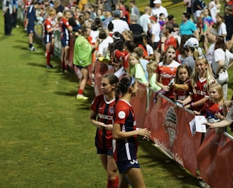 The image features a lively gathering of young soccer players and fans next to a field. Several female athletes in red and blue uniforms interact with an enthusiastic crowd standing behind a barrier. Some people in the crowd are holding objects, like pens and paper, possibly for autographs.