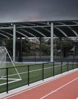 An outdoor sports facility featuring a running track and a soccer goal. The track is red, surrounded by a black metal fence, and the field is covered with artificial green turf. In the background, a basketball court is visible under a covered roof with metal supports. The sky is overcast, adding a moody atmosphere.