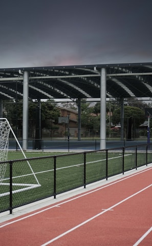 An outdoor sports facility featuring a running track and a soccer goal. The track is red, surrounded by a black metal fence, and the field is covered with artificial green turf. In the background, a basketball court is visible under a covered roof with metal supports. The sky is overcast, adding a moody atmosphere.