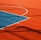 A vibrant sports court with sections of red and blue, featuring crisp white lines marking the boundaries. Shadows from a structure create geometric patterns across the surface, adding depth and interest to the scene.
