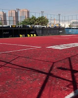 A sports court with a red playing surface and white lines, surrounded by a black mesh fence. In the background, tall buildings are visible, along with some trees and a few yellow chairs positioned near the fence.