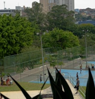 An outdoor recreational area in an urban setting featuring several sports courts and surrounded by lush green trees. People are engaged in various activities, including playing sports on the courts. In the background, there are tall buildings indicative of a cityscape.